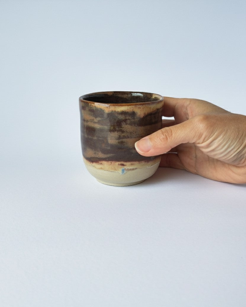 Hand holding a ceramic cup with a brown and beige glaze on a light gray background